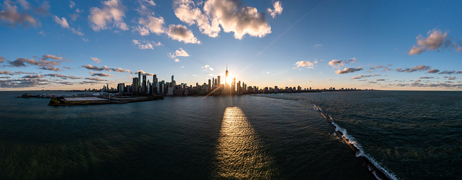 Wide angle aerial panorama of the Chicago skyline from Lake Michigan above the breakwater as the sun sets between highrise buildings casting a yellow glow on the water with blue sky and clouds above.