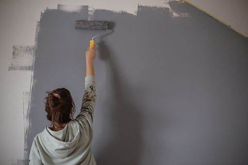 Young creative woman is painting interior wall. She is enjoying in renovating and refreshing her home.