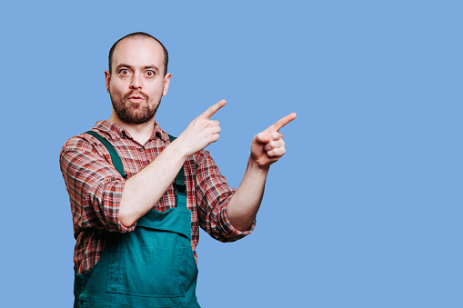 male workman, with a beard and an amazed expression on his face. He appears surprised as he points with his hand towards the empty space in from his left, looking at camera, studio shot, copy space