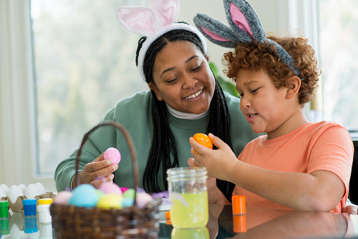 Beautiful mother and her Elementary age son are smiling and painting Easter eggs together at the kitchen table as part of their holiday traditions together. They are wearing costume rabbit ears on their heads.