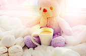 White cup and fluffy teddy bear toy on the window sill. Birthday or Valentines Day concept. Soft light and blurred effect