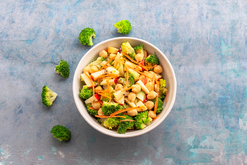 Broccoli salad add apple chickpeas in white bowl isolated on blue wood background close up, top view, healthy food concept.