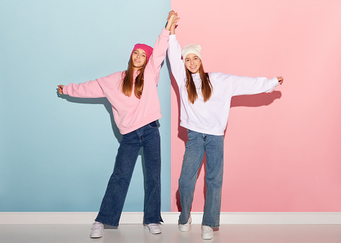 Friendship. Portrait of fashionable emotive teen girls in trendy warm clothes and hats having fun isolated over pink-blue background. Youth fashion, beauty, emotions, friendship and ad