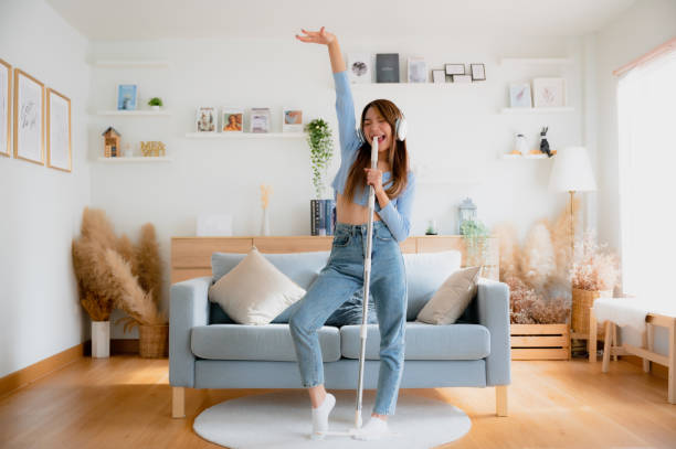 House cleaning with fun. Happy young asian housewife singing her favorite song during cleanup, using mop as microphone, enjoying domestic work. Young woman dancing and cleaning in living room stock photo