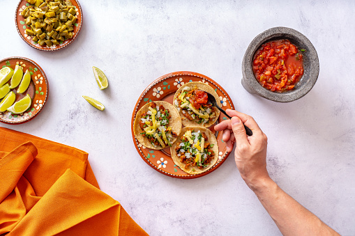Top view of crop anonymous person adding salsa sauce on taco served on colorful ornamental ceramic plate near sliced lime on white table