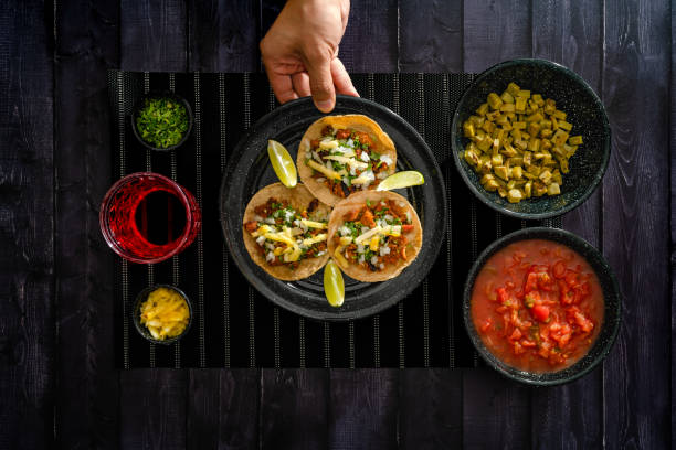 Mexican tacos served on black table near various toppings, top view of crop anonymous person serving tacos on black slate board with assortment of toppings and drink on table