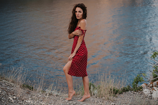 A young Caucasian woman poses in a red dress at the edge of a cliff near the beach and the sea.