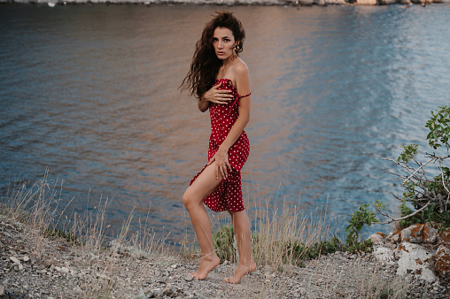 A young Caucasian woman poses in a red dress at the edge of a cliff near the beach and the sea.