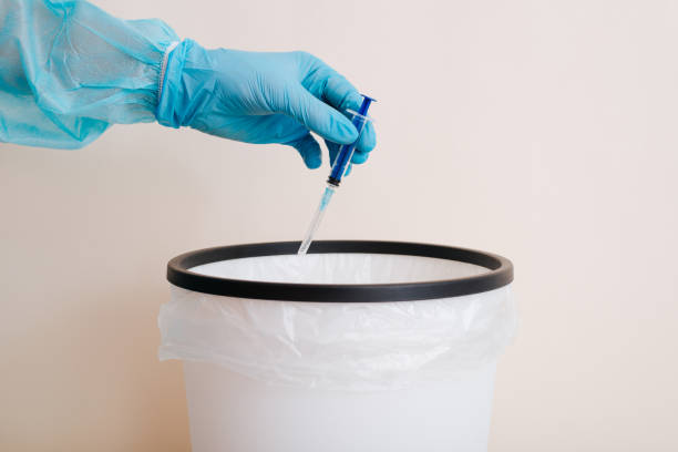 Doctor throwing used syringe into trash can, close-up of hand in protective medical glove stock photo