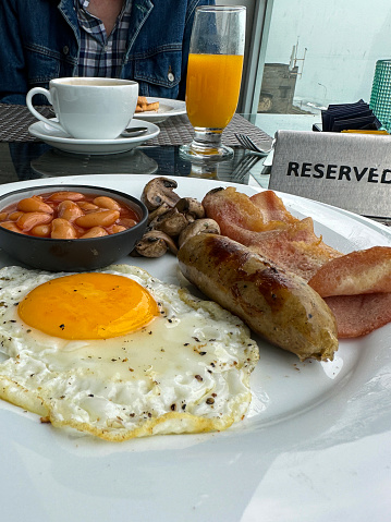 Stock photo showing close-up view of a white plate containing a Full English breakfast with bacon rashers, sausages, baked beans in a bowl, sunny side up fried egg and a pile of mushrooms.