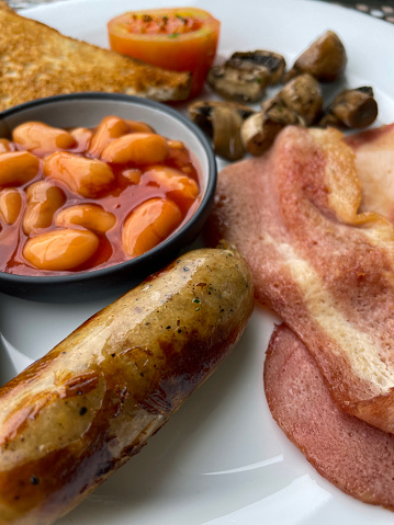 Stock photo showing close-up, elevated view of a white plate containing a Full English breakfast with bacon rashers, sausage, baked beans in a bowl, half a grilled tomato, pile of mushrooms and a slice of white bread toast.