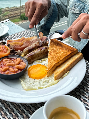 Stock photo showing close-up view of a white plate containing a Full English breakfast with bacon rashers, two sausages, baked beans in a bowl, half a grilled tomato, sunny side up fried egg, pile of mushrooms and white bread toast.