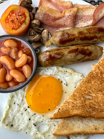 Stock photo showing close-up, elevated view of a white plate containing a Full English breakfast with bacon rashers, two sausages, baked beans in a bowl, half a grilled tomato, pile of mushrooms, sunny side up fried egg and a slice of white bread toast.
