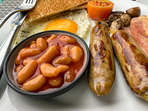 Stock photo showing close-up, elevated view of a white plate containing a Full English breakfast with bacon rashers, two sausages, baked beans in a bowl, half a grilled tomato, pile of mushrooms, sunny side up fried egg and a slices of white bread toast.