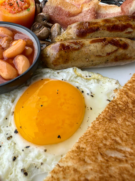 istock Full frame image of white plate containing Full English fried breakfast ingredients, two sausages, ramekin of baked beans, bacon rashers, tomato, mushrooms, fried egg, slice of toast on restaurant table, elevated view, focus on foreground 1459253852