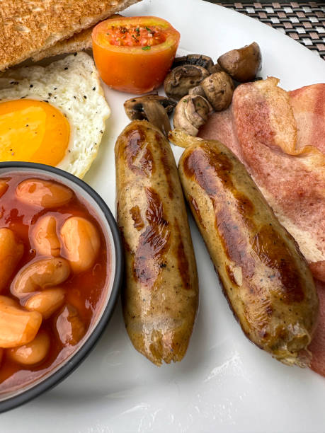 Stock photo showing close-up, elevated view of a white plate containing a Full English breakfast with bacon rashers, two sausages, baked beans in a bowl, half a grilled tomato, pile of mushrooms, sunny side up fried egg and a slice of white bread toast.
