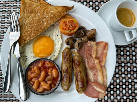 Stock photo showing close-up, elevated view of a white plate containing a Full English breakfast with bacon rashers, two sausages, baked beans in a bowl, half a grilled tomato, pile of mushrooms, sunny side up fried egg and a slices of white bread toast.