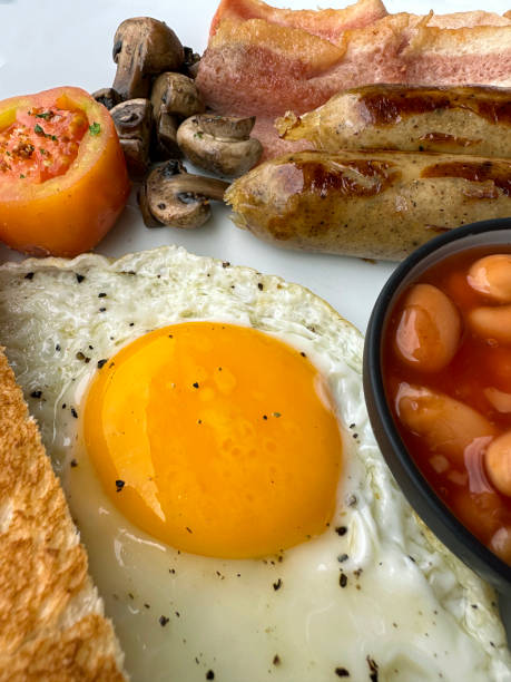 istock Full frame image of white plate containing Full English fried breakfast ingredients, two sausages, ramekin of baked beans, bacon rashers, tomato, mushrooms, fried egg, slice of toast, elevated view, focus on foreground 1459253860