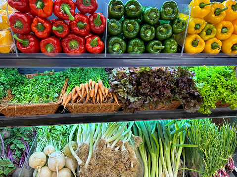 Stock photo showing a close-up view of a stacks of red, green and yellow bell peppers (Capsicum), beetroot, turnips, leeks, spring onions, coriander, carrots and lettuces being sold from a produce display at the fruit and vegetable grocery section of a supermarket.
