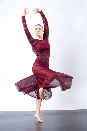 Young woman dancing in the studio in a burgundy dress against a white background, turning on one foot, with arms raised and dress billowing.