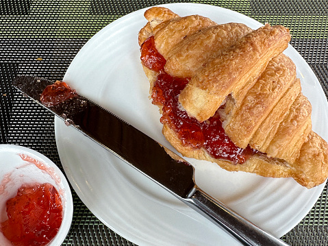 Stock photo showing a close-up, elevated view of a meal with golden brown croissant, french pastry treats served with strawberry jelly preserve in a white bowl, a delicious breakfast brunch.