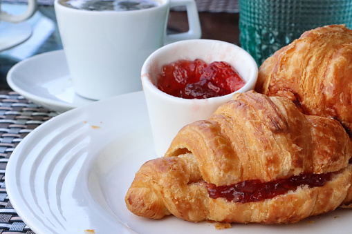 Stock photo showing a close-up view of a meal with two golden brown croissants, french pastry treats served with strawberry jelly preserve in a white bowl, a delicious breakfast brunch.
