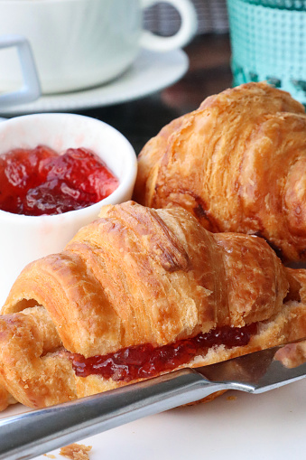 Stock photo showing a close-up view of a meal with two golden brown croissants, french pastry treats served with strawberry jelly preserve in a white bowl, a delicious breakfast brunch.