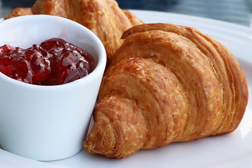 Close up stock photo depicting a breakfast meal with two golden brown croissants, french pastry treats served with strawberry jam / jelly preserve in a white cup, a delicious brunch