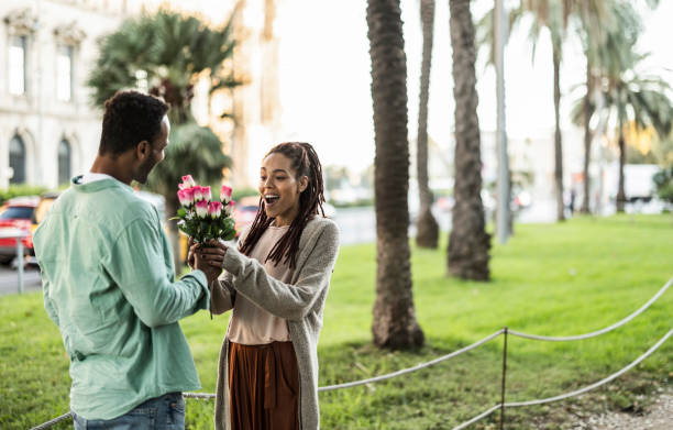 l'uomo romantico dà un mazzo di rose alla sua ragazza per strada - lei è sorpresa e felice - - men african descent giving flower foto e immagini stock