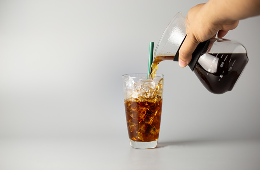 Hand pouring black coffee into ice glass from pitcher. cold coffee beverage on grey background.