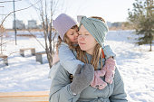 A girl hugs her mother while sitting on a bench in the park in winter