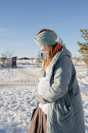 A happy pregnant young woman with her eyes closed, hugging her stomach in the park in winter, in a warm down jacket.