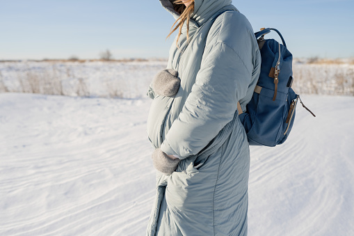 A pregnant young woman with a backpack hugs her stomach with warm mittens while standing in nature in winter.