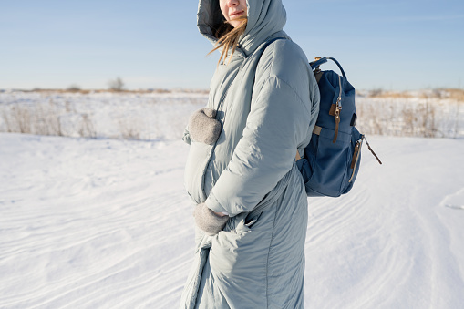 A pregnant young woman with a backpack hugs her stomach with warm mittens while standing in nature in winter.