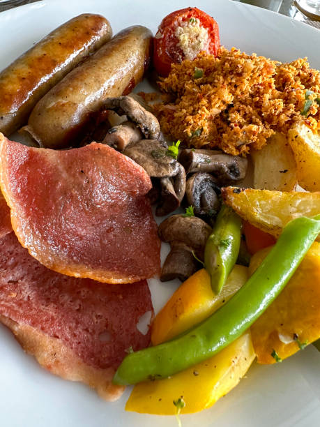 istock Full frame image of white plate containing fried breakfast ingredients, two sausages, bacon rashers, grilled tomato, mushrooms, coconut samba, potato wedges on restaurant table, elevated view, focus on foreground 1459274977