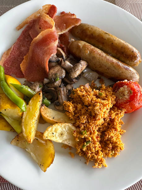 istock Close-up image of white plate containing fried breakfast ingredients, two sausages, bacon rashers, grilled tomato, mushrooms, coconut samba, potato wedges on restaurant table, elevated view 1459274981