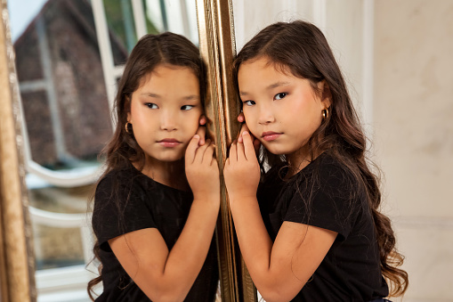 Asian little kid fashion model posing at mirror indoors, looking at camera. Close up portrait stylish young lady child girl actress in living room. Fashionable concept. Copy text space for advertising