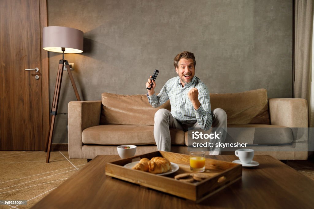 Cheerful man celebrating while watching a sports game on TV. Cheerful man having fun while celebrating the success of his sports team while watching a TV at home and looking at camera. Copy space. Television Set Stock Photo