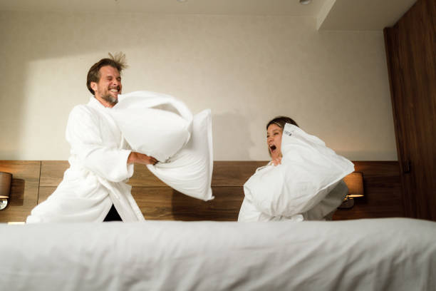 pareja alegre teniendo una pelea de almohadas en una cama. - bed couple pillow fight men fotografías e imágenes de stock