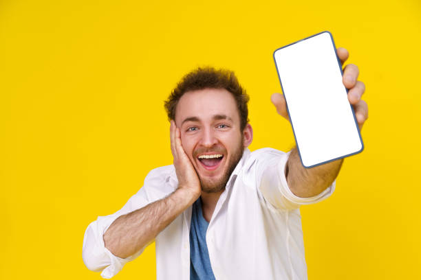 istock Great offer. Young happy man holding smartphone showing a white empty screen and exciting to win isolated over yellow background, celebrating success. Product placement. Empty space or copy space 1459280222