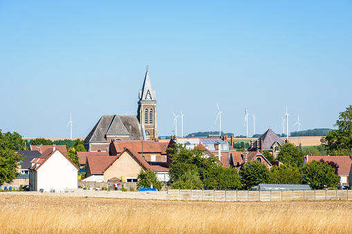 View over the small rural village of Vendeuil, France, and its steeple, surrounded by cultivated fields, with the wind turbines of the Villes dOyses wind farm in the background.
