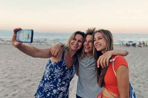 Mother and teenagers walking on beautiful beach. They are hugging and taking selfies. \nCanon R5