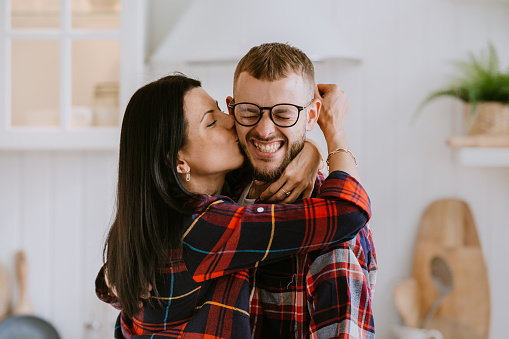 Happy couple celebrating success at home. Young Italian man in glasses eyes closed excited, winner. Brunette hisp[anic girl kissing husband sad him that she is pregnant. Happy family. Triumph.