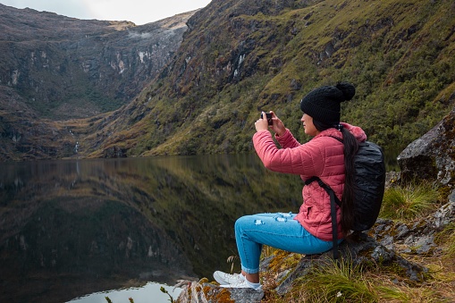 A girl in a pink jacket with a backpack sitting on a stone by the lake and taking a photo on the phone in the mountains