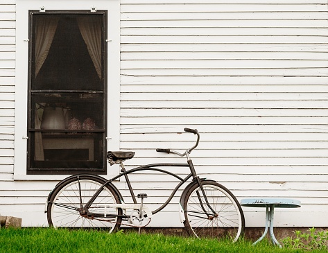 A closeup shot of a retro bicycle on a green lawn leaning on a white wooden wall with a long window