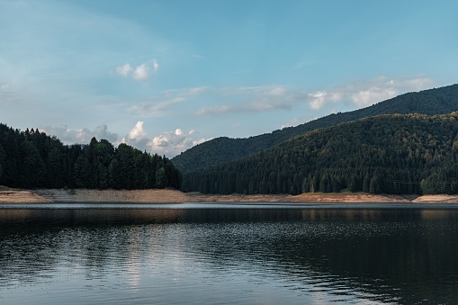 A landscape of a lake surrounded by dense green mountain forests under blue cloudy sky