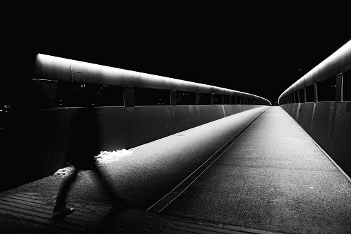 A grayscale shot of the person walking along the bridge at night.