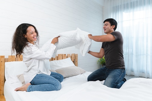A happy Thai couple fighting by pillows on a bed and having fun together at home