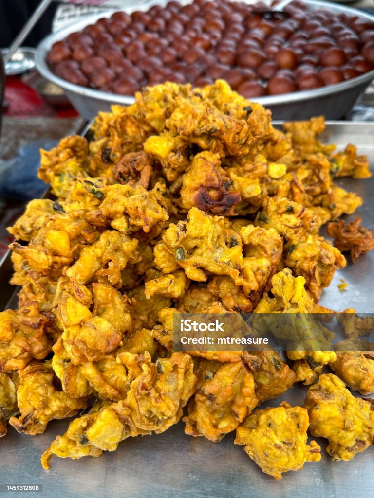 Close-up image of pile of crispy onion bhajis on rectangular catering tray, Indian street cuisine vegetarian snack, stainless steel bowl of Gulab jamun (Rose water berry) Mithai (sweets) in background, elevated view, focus on foreground Stock photo showing a close-up view of a rectangular, stainless steel tray containing a heap of crispy, deep fried onion bahjees a popular street food. Besides these treat is a bowl containing Indian sweets called Gulab jamun, made from khoya (dried evaporated milk solids) dough rolled, deep fried and then soaked in a sugar syrup (water, sugar cardamom, rosewater, and saffron).
These candies are the national dessert of India. Fried Stock Photo