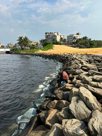 Stock photo showing close-up view of fisherman with long fishing rod sitting on a natural rocky sea defence stack of large rocks and stones, which have been piled up to provide a sea defence on a sandy beach in Colombo, Sri Lanka. This 'riprap' rock armour is helping to stop fine sand from being washed away due to gradual coastal erosion.